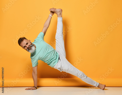 Smiling bearded grey-haired mature man practicing yoga lesson studio shot. Handsome brutal male person exercising side bar with twine indoor full length portrait over yellow copy space photo