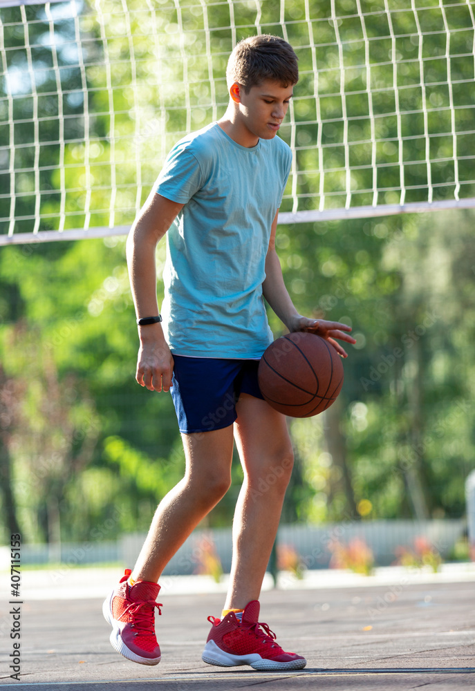 Cute young boy plays basketball on street playground in summer. Teenager in green t shirt with orange basketball ball outside. Hobby, active lifestyle, sport activity for kids. Cute young boy plays basketball on street playground in summer. Teenager in green t shirt with orange basketball ball outside. Hobby, active lifestyle, sport activity for kids.