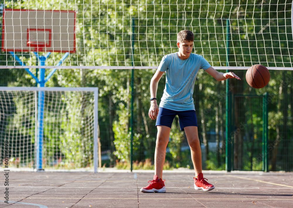 Cute young boy plays basketball on street playground in summer. Teenager in green t shirt with orange basketball ball outside. Hobby, active lifestyle, sport activity for kids.	