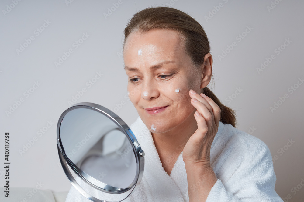 Mature woman in a white morning gown looking in the mirror. Home skin care for older skin. Mature woman in a white morning gown looking in the mirror. Home skin care for older skin.