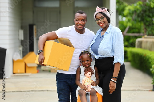 African American family is moving in to a new house with father holding cardboard box along with suitcase and baggage with happy smile