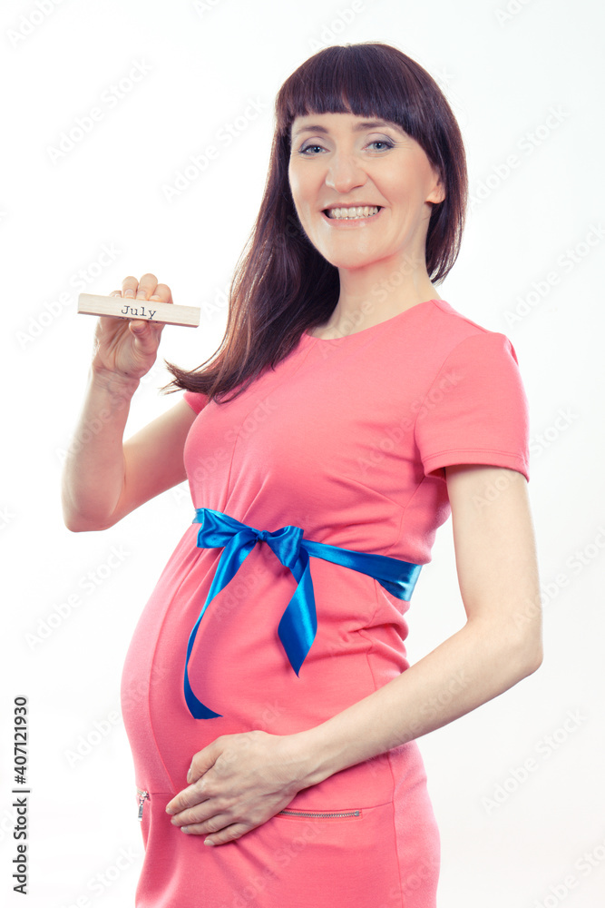 Happy smiling woman in pregnant wearing pink dress with blue ribbon and showing word july. Expecting for newborn and expansion of family Happy smiling woman in pregnant wearing pink dress with blue ribbon and showing word july. Expecting for newborn and expansion of family