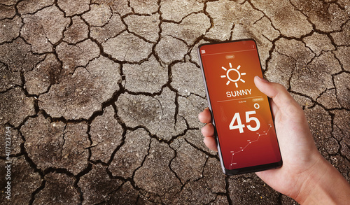 Top View,Hand holding smart phone with weather forecast High Temperature Weather Hot Sunny Day on screen and dry cracked earth background.