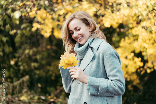 Charming cheerful young woman in an autumn park landscape with a bouquet of yellow leaves. Portrait of a lady in a gray coat.
