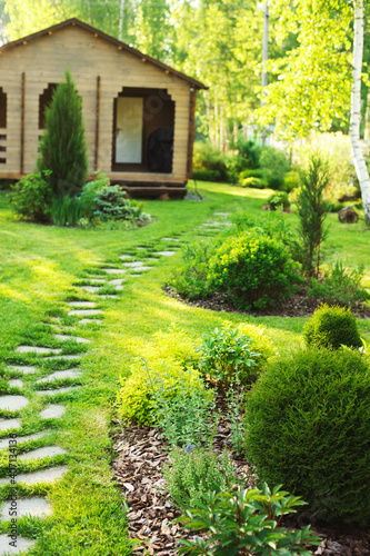 stone pathway in summer garden with beautiful wooden house on background. Landspace design with conifers and shrubs
