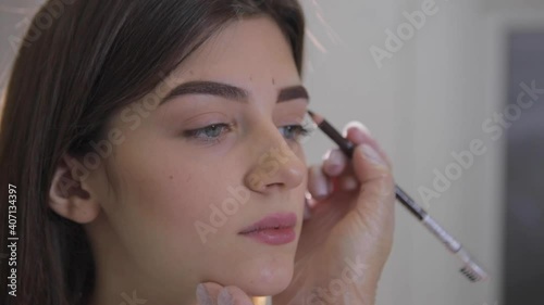 Beautician applies elegant makeup on brows of attractive young woman with cosmetic pencil in beauty salon closeup slow motion