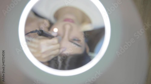 Master applies permanent makeup on young woman eyebrows with machine in salon upper view through magnifier glass slow motion