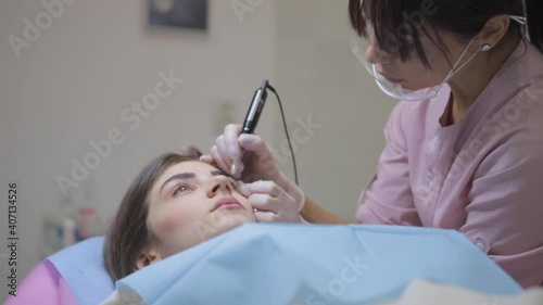 Concentrated woman in pink uniform applies permanent makeup on pretty client brows with tattoo machine in salon slow motion