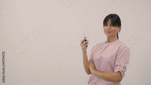 Cheerful woman beautician in pink uniform with contemporary tattoo machine poses for camera on beige background slow motion
