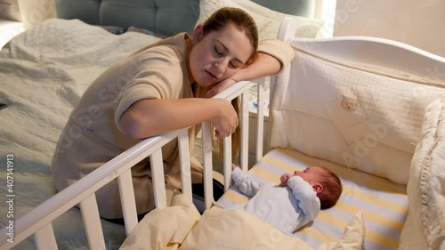 Young tired and exhausted mother fallen asleep while rocking crib of her newborn baby at night. Concept of sleepless nights and parent depression after childbirth