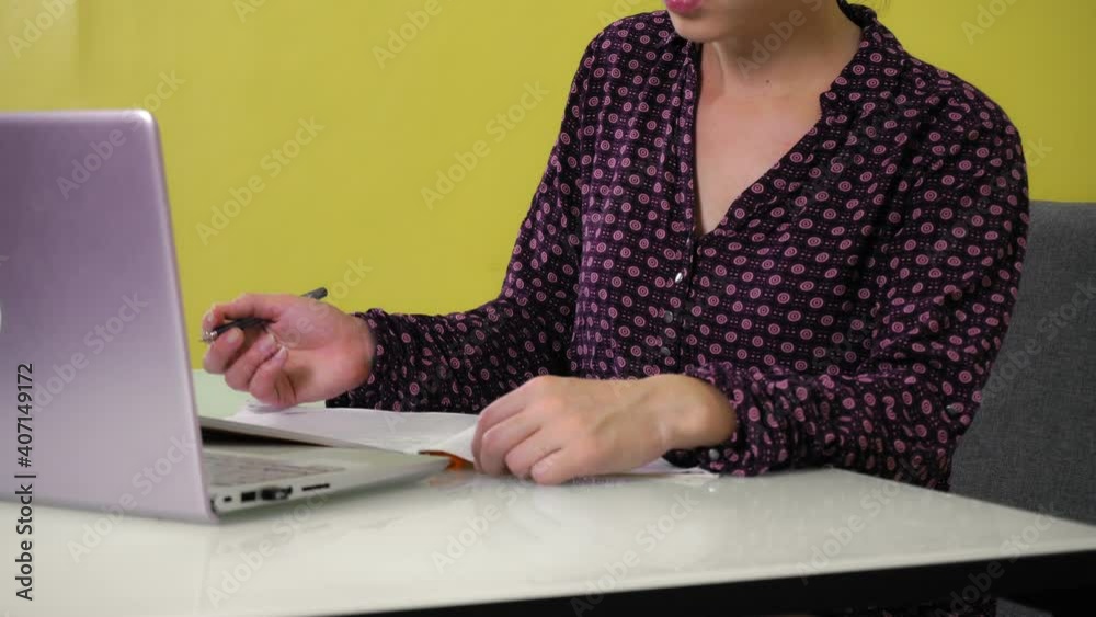 A young mixed race woman sits at home and is engaged online on a laptop, making notes in a notebook. A woman communicates at work or in a distance class. 