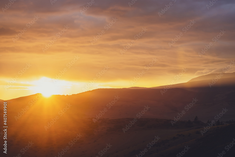 Atmospheric landscape with silhouettes of mountains with trees on background of dawn sky with sun circle and orange sun rays. Colorful nature scenery with sunset or sunrise of illuminating color.
