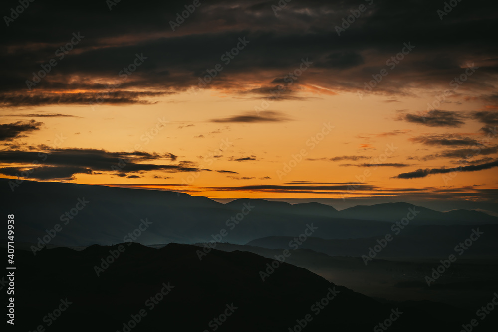 Scenic dawn mountain landscape with golden low clouds in valley among dark mountains silhouettes under sunset or sunrise sky. Vivid scenery with low clouds in mountain valley in illuminating color.