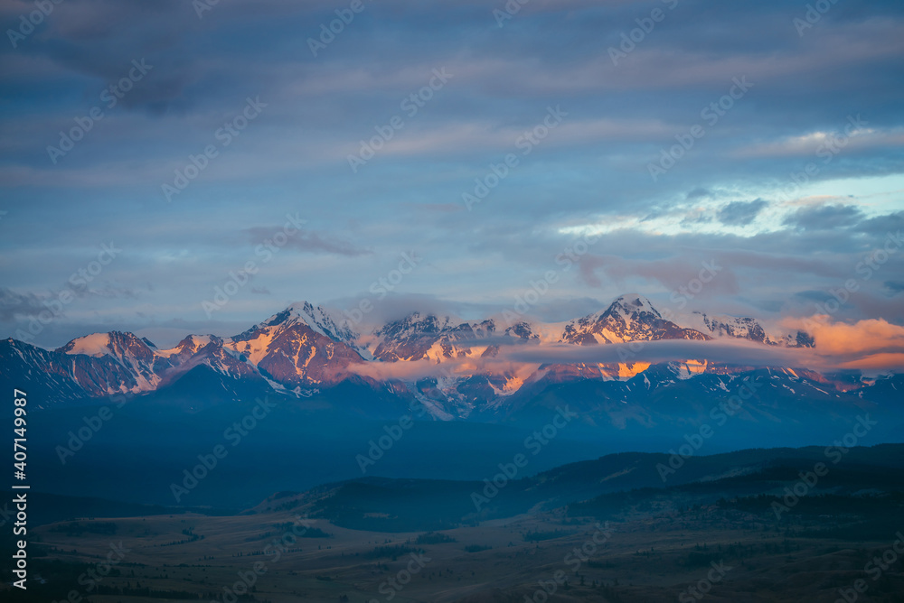 Obraz premium Scenic mountain landscape with great snowy mountain range lit by orange dawn sun among low clouds. Awesome alpine scenery with snowy high mountain ridge under blue cloudy sky at sunset or at sunrise.