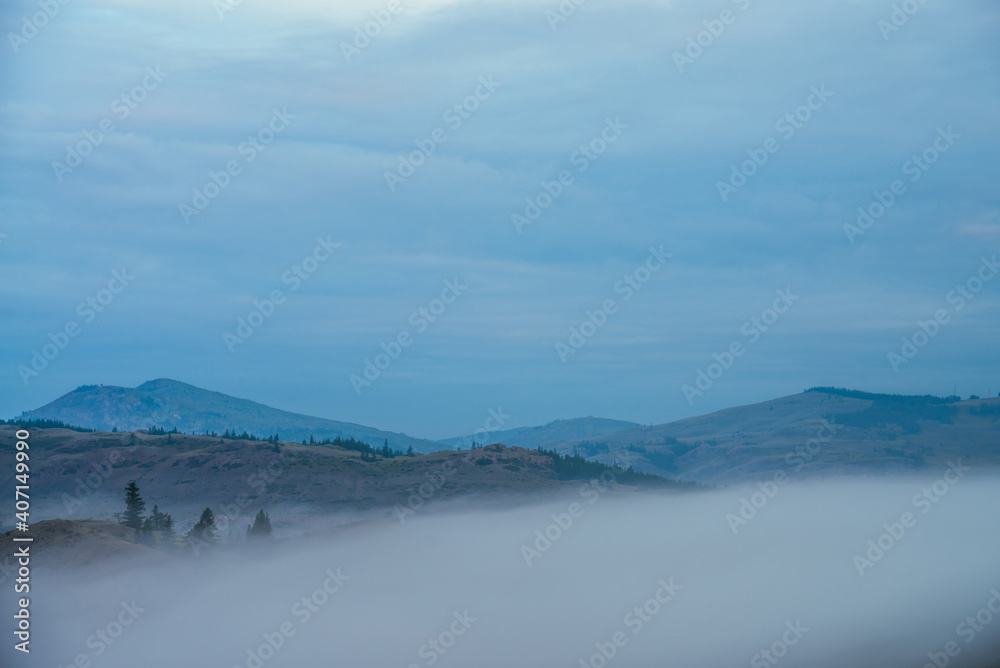 Atmospheric mountain landscape with green hills above clouds and blue cloudy sky. Minimal alpine scenery with big low cloud in mountains. Dense fog under hills with trees. Early morning in mountains.