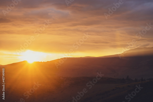 Atmospheric landscape with silhouettes of mountains with trees on background of dawn sky with sun circle and orange sun rays. Colorful nature scenery with sunset or sunrise of illuminating color. © Daniil