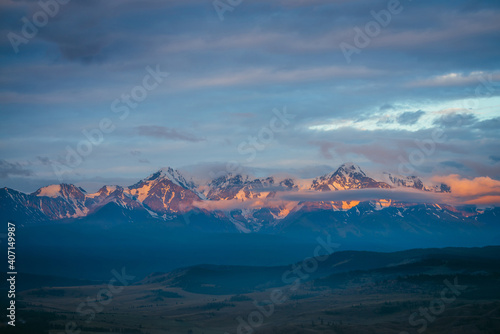 Scenic mountain landscape with great snowy mountain range lit by orange dawn sun among low clouds. Awesome alpine scenery with snowy high mountain ridge under blue cloudy sky at sunset or at sunrise.