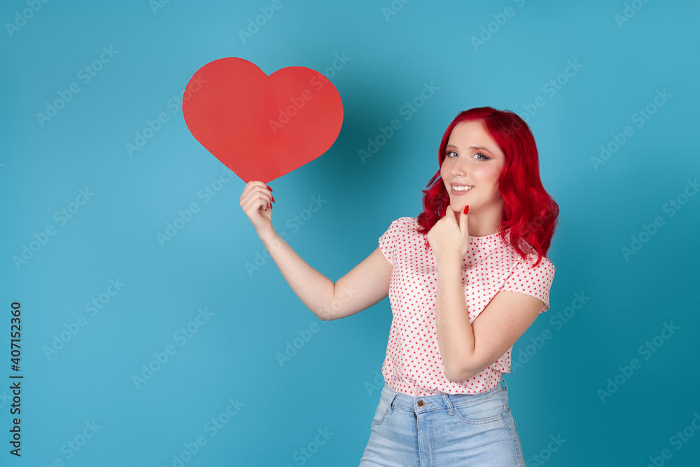 smiling beautiful young woman with red hair holding red paper heart and scratching her chin isolated on blue background. smiling beautiful young woman with red hair holding red paper heart and scratching her chin isolated on blue background.