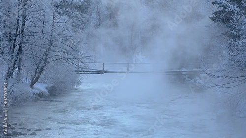 Pristine cold Alpine river flowing under small suspension bridge in winter. Snow covers river bank in Slovenia. Water evaporating into mist above stream. Colorful blue and pristine creek