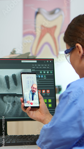 Nurse having video call with expert stomatologic medic while doctor is working with patient in background. Stomatologist assistant listening dentist using mobile webcam sitting on stomatological chair