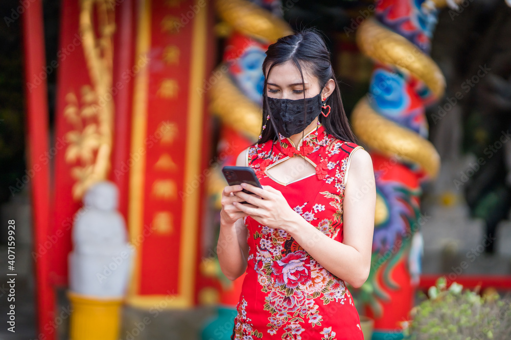 Asian young woman wearing red traditional Chinese cheongsam decoration and wear protective mask and smartphone for Chinese New Year Festival at shrine,Prevention of the spread of COVID-19 virus