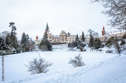 Pruhonice, Czech Republic - January 16 2021: View of the castle and the Church of the Birth of the Virgin Mary over a lake, all covered with white snow. Grey sky in the background.