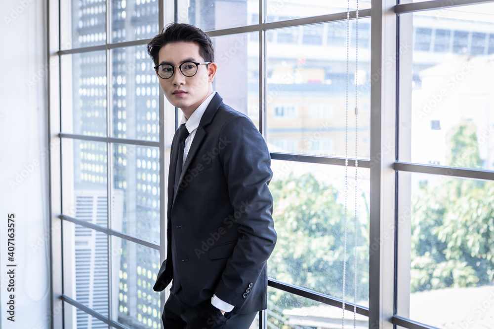 Portrait of successful feeling winner handsome young asian businessman with glasses wear a business suit of man in blue jacket and blue shirt looking at the window In the office room background. Portrait of successful feeling winner handsome young asian businessman with glasses wear a business suit of man in blue jacket and blue shirt looking at the window In the office room background.