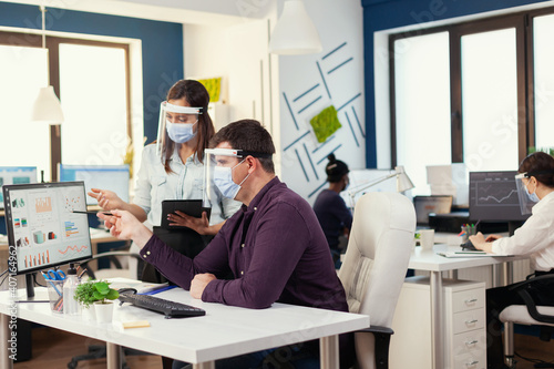 Employess working together wearing face mask as safety precaution during covid19. Multiethnic team in new normal financial office in corporate company typing on computer, taking notes on tablet.