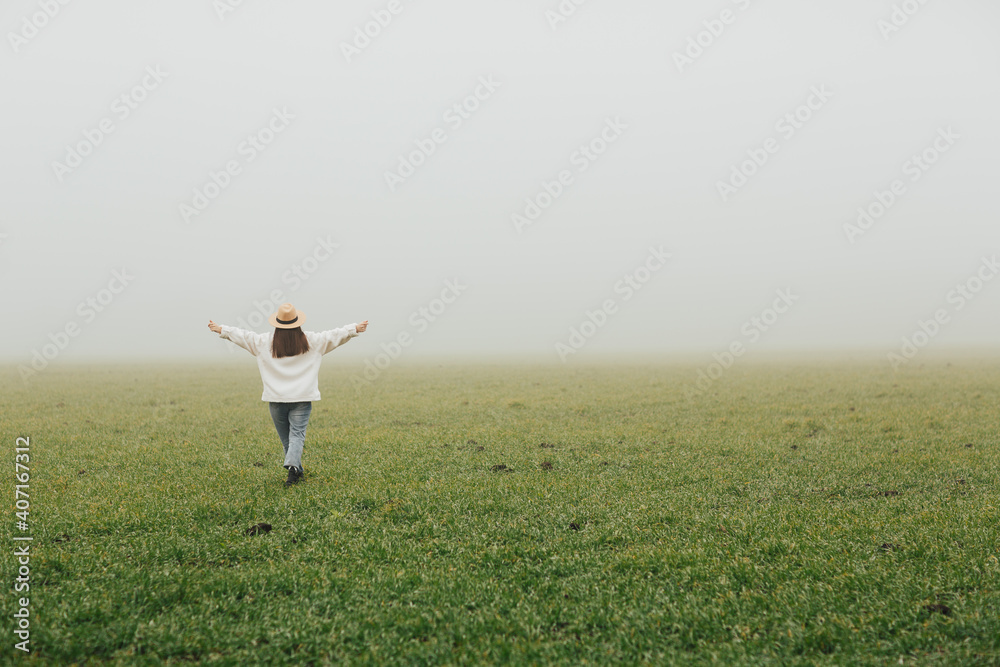 Happy girl standing with back raise her hands up on green field in the foggy morning.