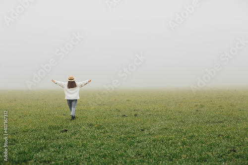 Obraz na płótnie Happy girl standing with back raise her hands up on green field in the foggy morning. w salonie