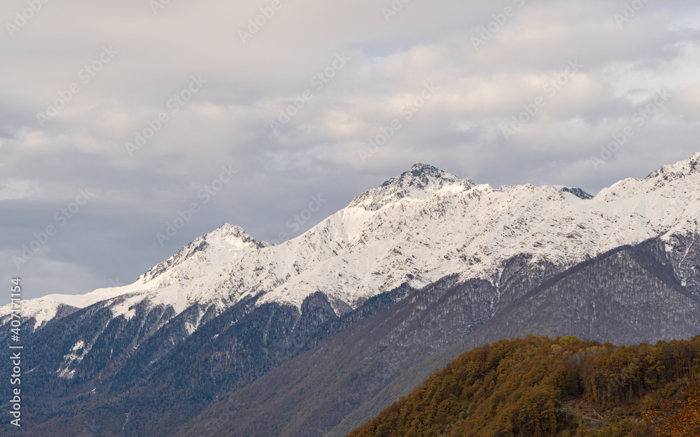 Snow-capped peaks of Caucasus Mountains from observation deck in resort village of Krasnaya Polyana. Mountain peaks and autumn forest on clear sunny day against backdrop of November cloudy sky. Snow-capped peaks of Caucasus Mountains from observation deck in resort village of Krasnaya Polyana. Mountain peaks and autumn forest on clear sunny day against backdrop of November cloudy sky.