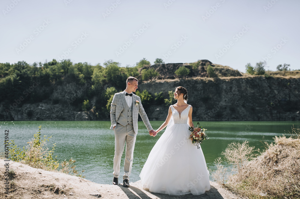 Stylish groom in a gray tailcoat with a black bow tie and a beautiful, curly-haired bride in a white long dress are standing holding hands near the river and rocks. Wedding portrait of newlyweds.