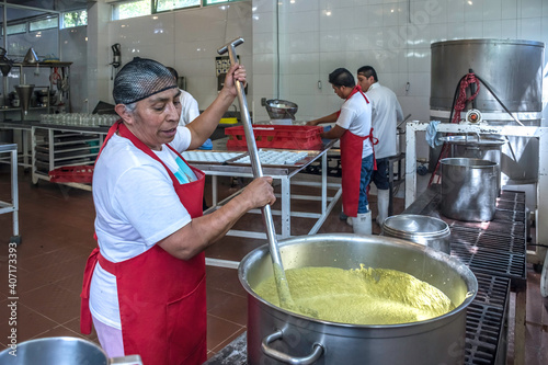 Latin woman kitchen worker stirring sauce in large industrial cooking pot.