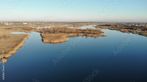 The estuary of a river with blue water. On the bank and in the middle of the river there are dry grass and reeds. There are village with small residential houses in the distance
