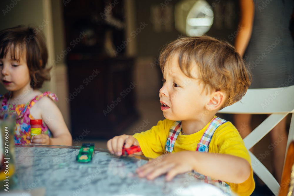Little child boy be capricious because want to sleep. Kid cry while sit at table in home with his small sister on backgriund. Little child boy be capricious because want to sleep. Kid cry while sit at table in home with his small sister on backgriund.