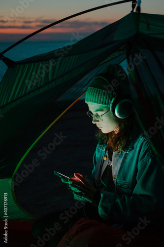 Young woman, millennial teenager or generation z sit in tent and use smartphone. Woman listen to headphones music or podcast in streaming app or online. Blue hour cinematic photo of solitude travel