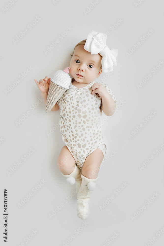 Fototapeta premium Cute baby with knitted ice cream. Adorable stylish baby in stylish clothes and headband looking at camera while lying on gray background. Fashion baby.