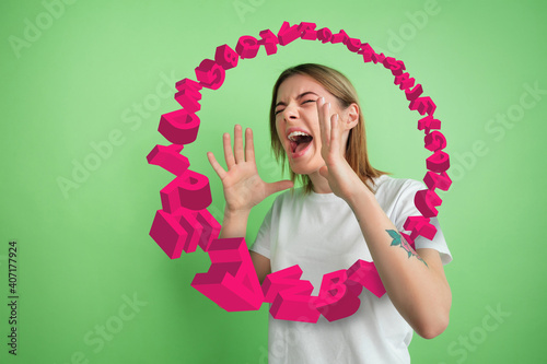 Woman shouting, screaming angry, highly tensioned on green studio background. Sales, offer, business, cheering fun concept. Stream of letters flying from the inside of rupor. Copyspace for ad. photo