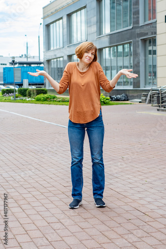 An adult redhead woman in casual clothes stands in the street spreading her arms and shrugging her shoulders in a questioning gesture. She does not know what is happening and what will happen next.