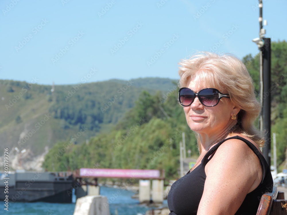 Happy blonde mature woman with sunglasses sitting outdoor and thinking. Senior woman relaxing on  bench  in a summer day. Portrait of middle aged woman smiling and daydreaming.