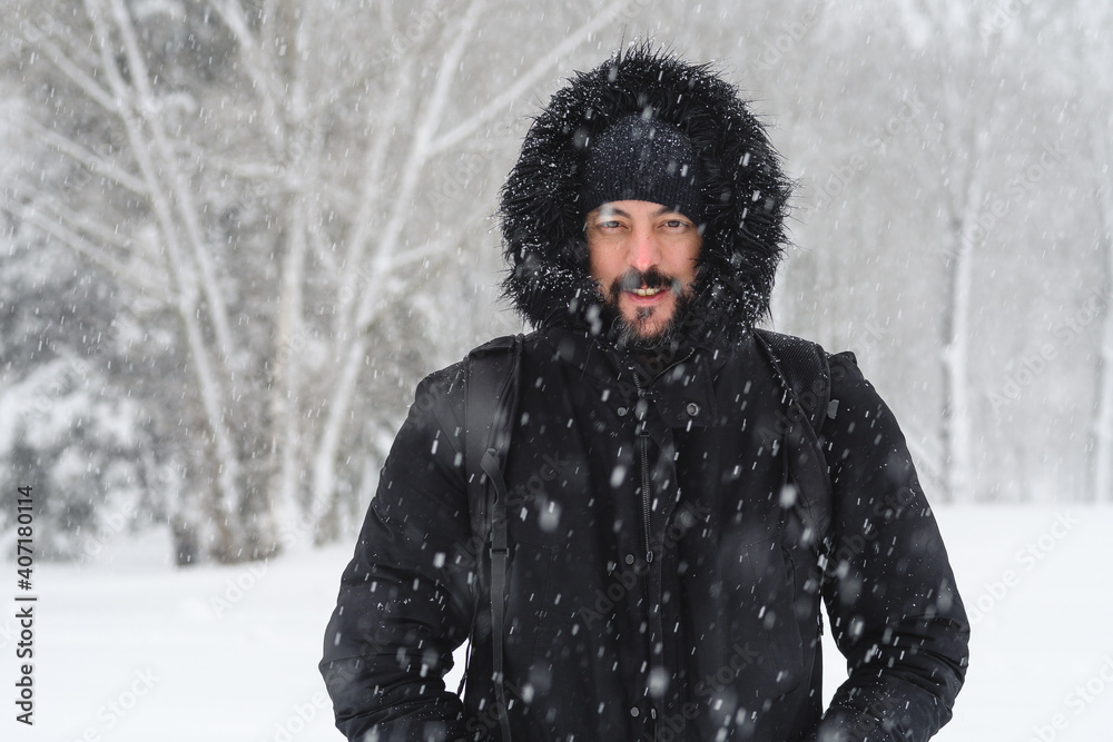 portrait of a young hipster man with a beard wearing a coat and hat from the cold while it is snowing in winter in a park with snowy trees in the background portrait of a young hipster man with a beard wearing a coat and hat from the cold while it is snowing in winter in a park with snowy trees in the background