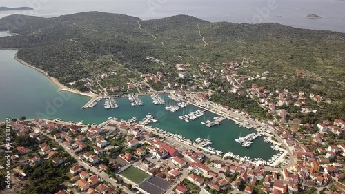 A descending drone shot over a gorgeous marina during daytime. A yacht is gliding over clear blue water, pulling out of the bay, surrounded by green trees and medeteranian houses. photo