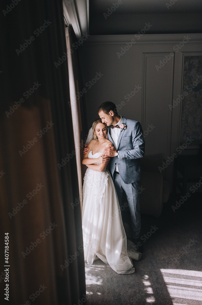 Fototapeta premium Wedding portrait of stylish groom in gray plaid suit and cute blonde bride with curly hair near the window with curtains in the hotel room. Interior photography.