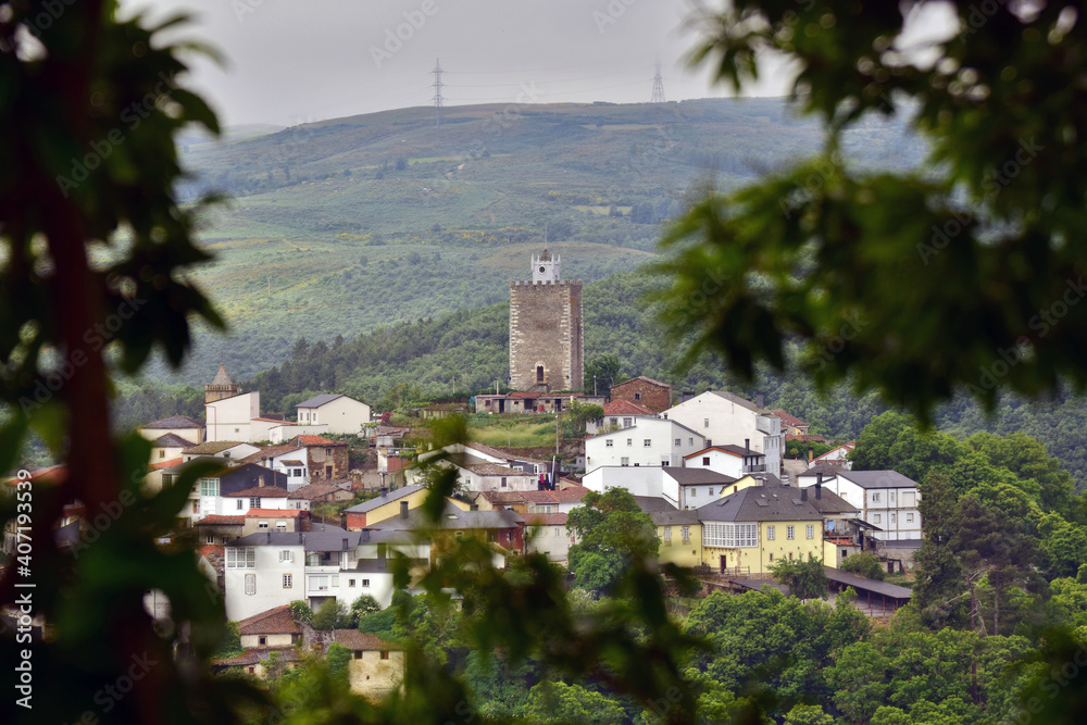 Rural landscape of a mountain village in Galicia. In the image you can see several houses, a tower and the bell tower of a church. In the background the sky and mountains with electric towers