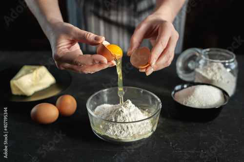 The process of preparing the dough. The woman breaks the egg and adds it to a bowl of flour. In women's hands an egg and a knife. Ingredients for the dough on a dark background.