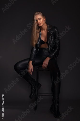 Beautiful sexy blonde girl in black leather clothes posing while sitting on a high stool on a dark gray background