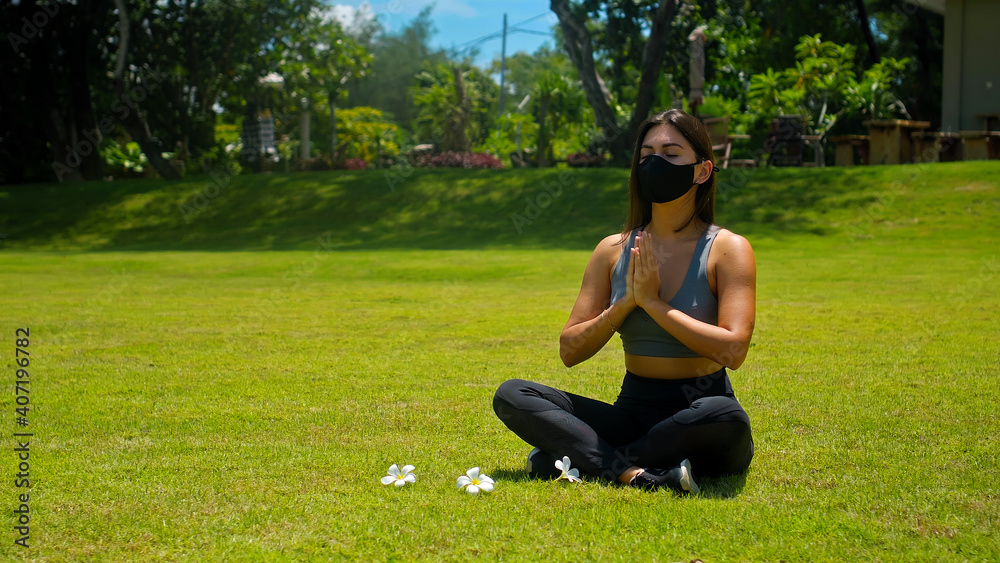Young European brunette girl practices alone yoga in nature sitting on green grass in lotus pose wearing a black protective mask