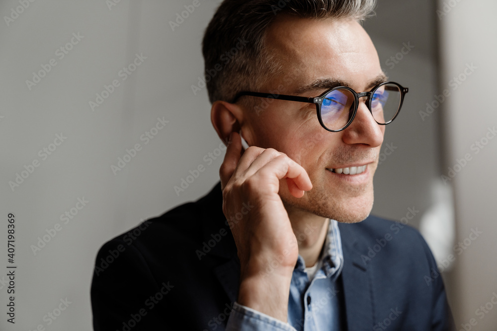 Pleased grey man smiling and using wireless earphones while working