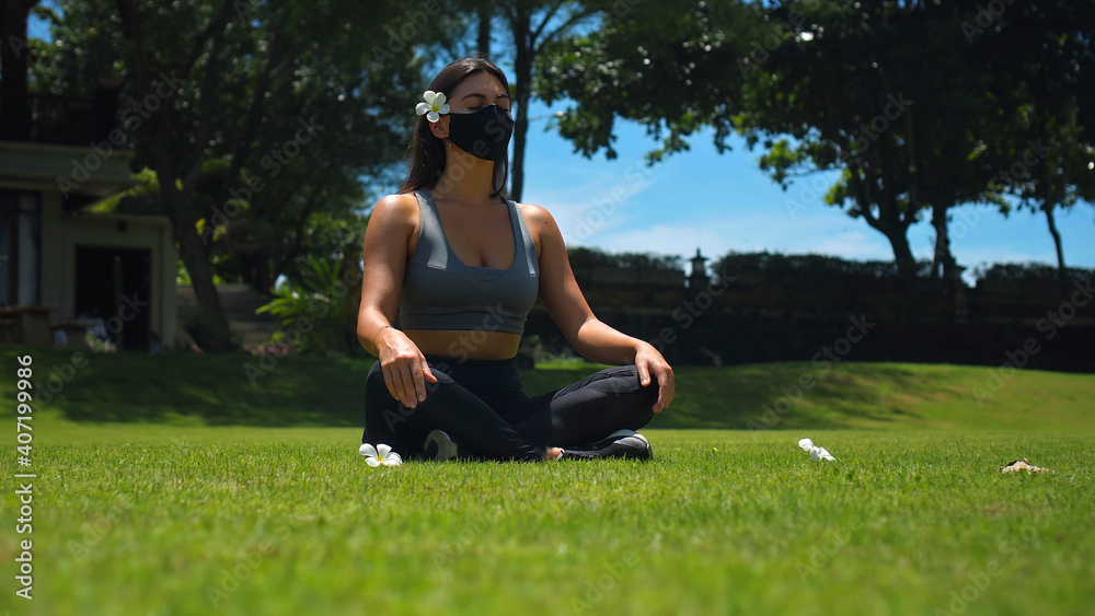 Fototapeta premium Young European brunette girl practices alone yoga in nature sitting on green grass in lotus pose wearing a black protective mask