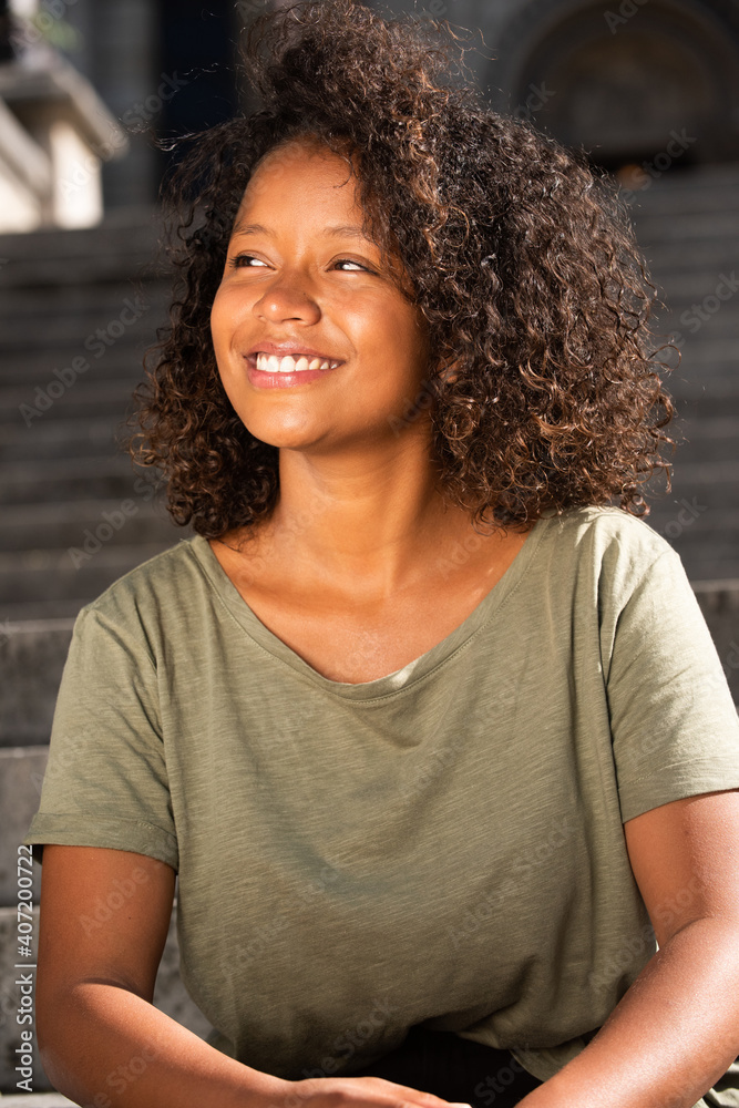 Fototapeta premium Portrait happy young woman with curly hair sitting outside looking away